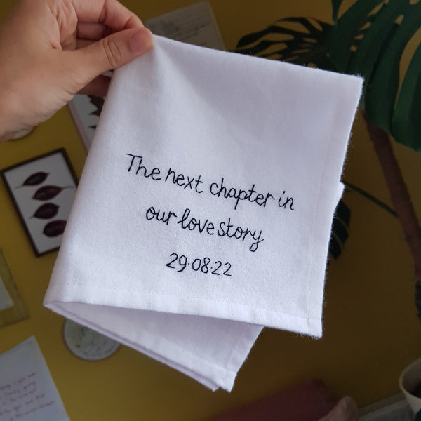 A white cotton handkerchief that is embroidered in black thread with the message ‘The next chapter in our love story 29.08.22'.