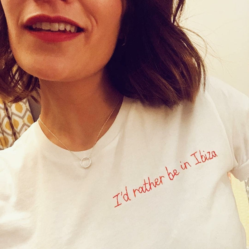 A white woman is wearing a white shirt with 'I'd rather be in Ibiza' embroidered on.