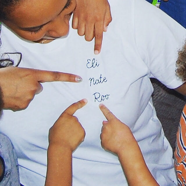 A mum is wearing a white t-shirt with the names of her children embroidered on it - her children are all pointing at them.
