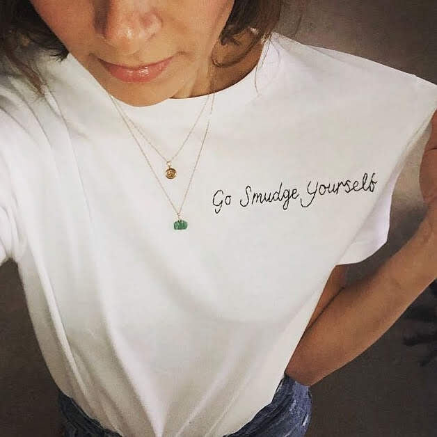 A white woman is wearing a white cotton t-shirt with 'Go Smudge Yourself' embroidered on it in black thread.