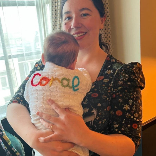 Woman holding a baby wearing a baby cardigan with 'Coral' embroidered on it in colourful threads.