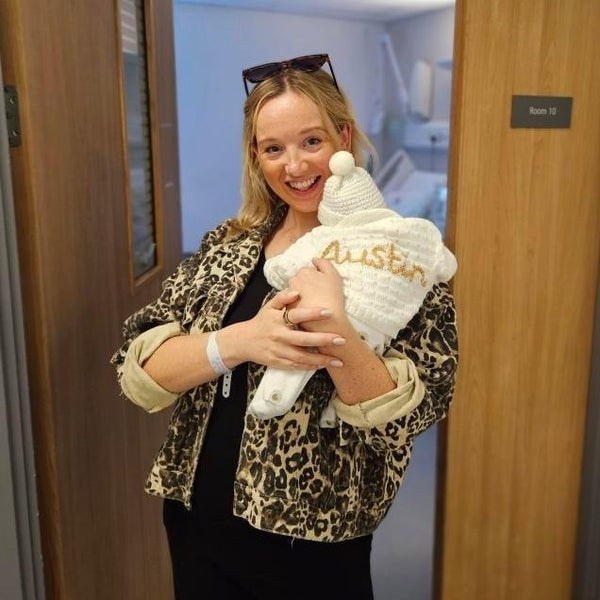 Woman holding a baby wearing a white knitted cardigan with 'Austin' embroidered on it, standing in a hospital doorway.