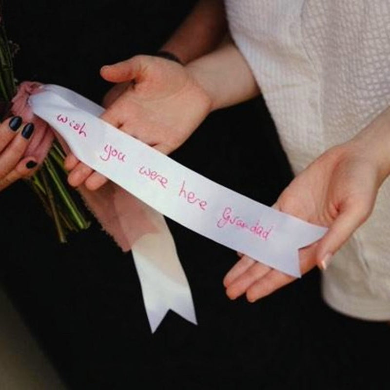 A white satin ribbon is tied around a wedding bouquet of flowers. The ribbon is held in the bride's hands. There is a handwritten message embroidered on the ribbon that read 'wish you were here Grandad' in pink thread.