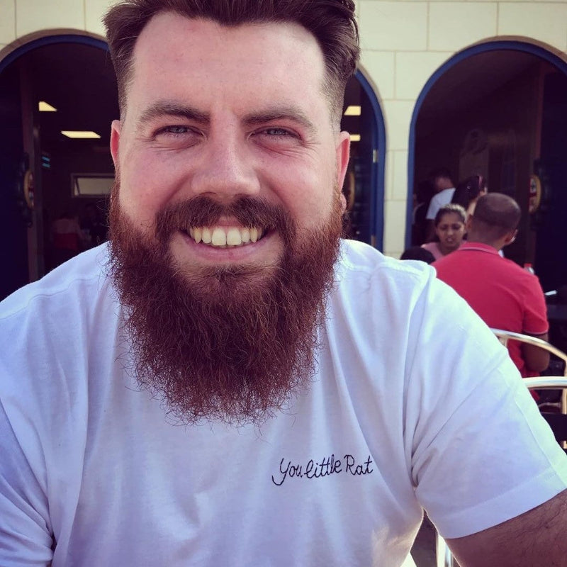 Man with a beard wearing a white t-shirt that is embroidered with the phrase 'You little Rat'.