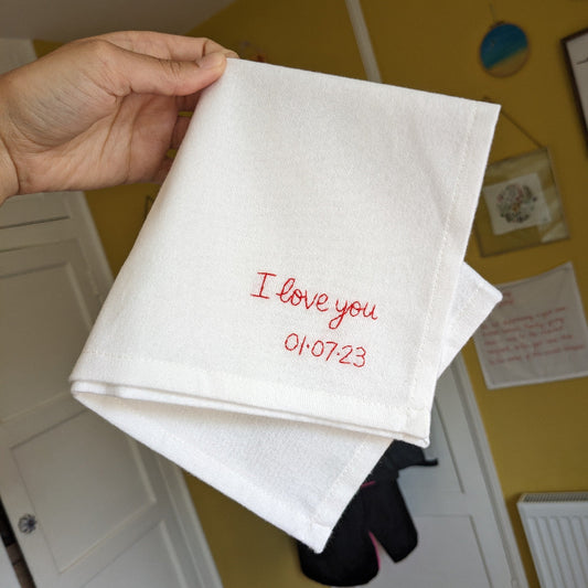 A white cotton handkerchief that is embroidered in red thread with the message ‘I love you 01.07.23'.