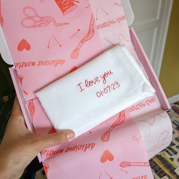 A white cotton handkerchief that is embroidered in red thread with the message ‘I love you 01.07.23'.