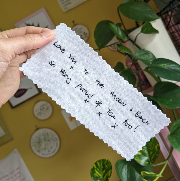 A white cotton wedding dress label with black embroidered handwriting that reads 'Love you to the moon + back + so very proud of you too! Xxx'