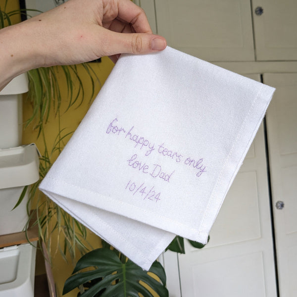 A white cotton handkerchief that is embroidered in lavender thread with the message ‘for happy tears only, love Dad 10/4/24'.