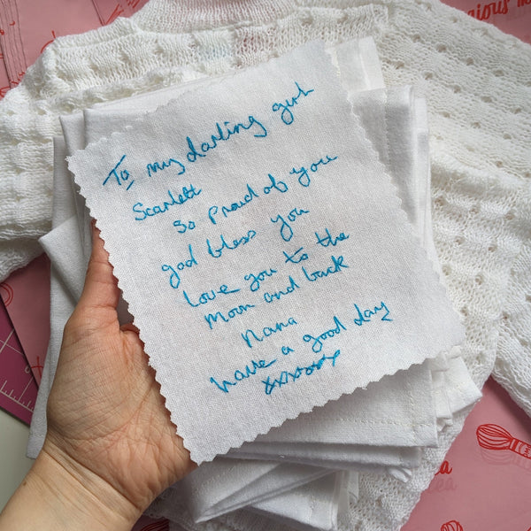 A white cotton wedding dress label with blue embroidered handwriting that reads 'To my darling girl Scarlett, so proud of you, god bless you, love you to the moon and back. Nana. Have a good day xxxxx'