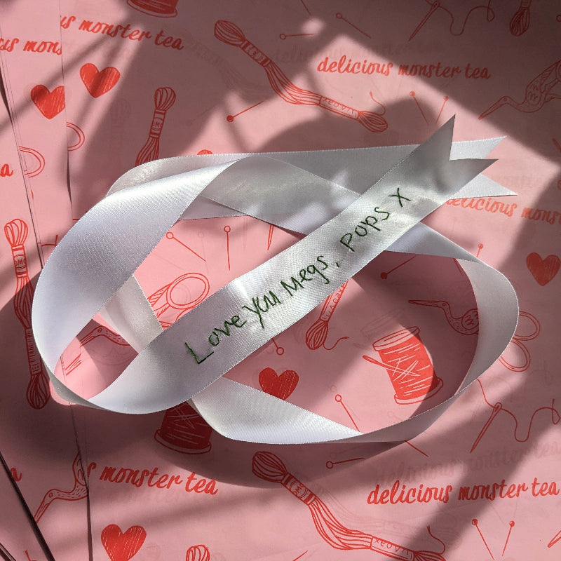 White ribbon with green handwriting embroidered on it. The embroidery reads 'Love you Megs, Pops x.
