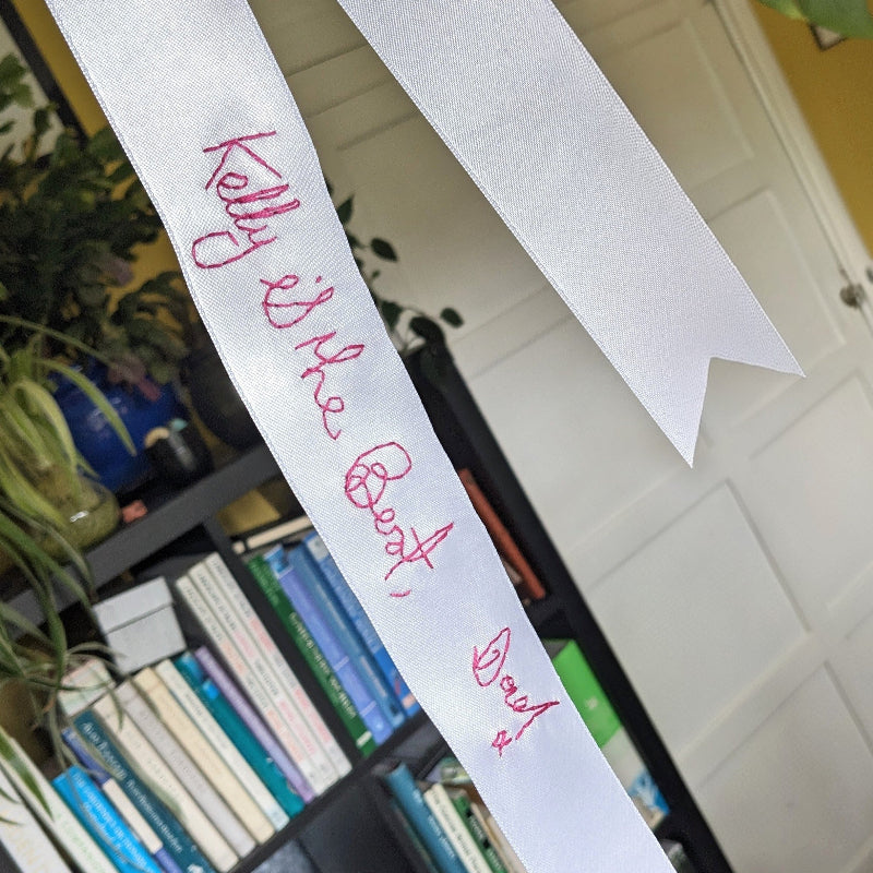 White ribbon with pink handwriting embroidered on it. The embroidery reads 'Kelly is the best, Dad x'.