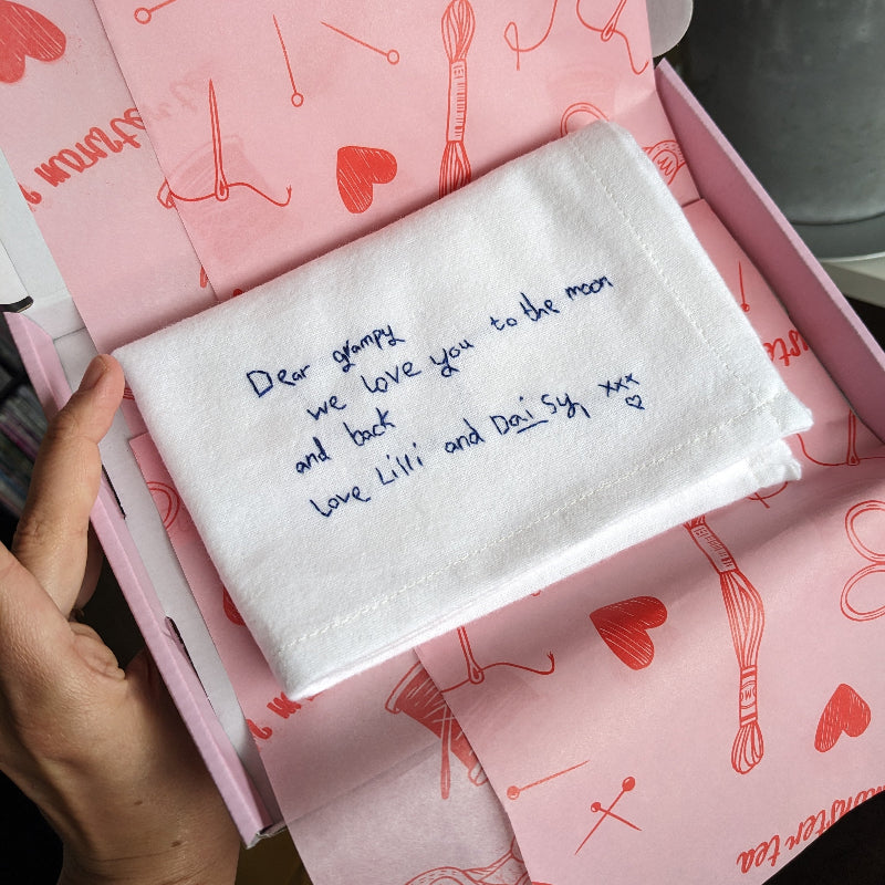 A white cotton handkerchief with navy embroidered kid's handwriting that reads 'Dear grampy, we love you to the moon and back. Live Lilli and Daisy xxx'