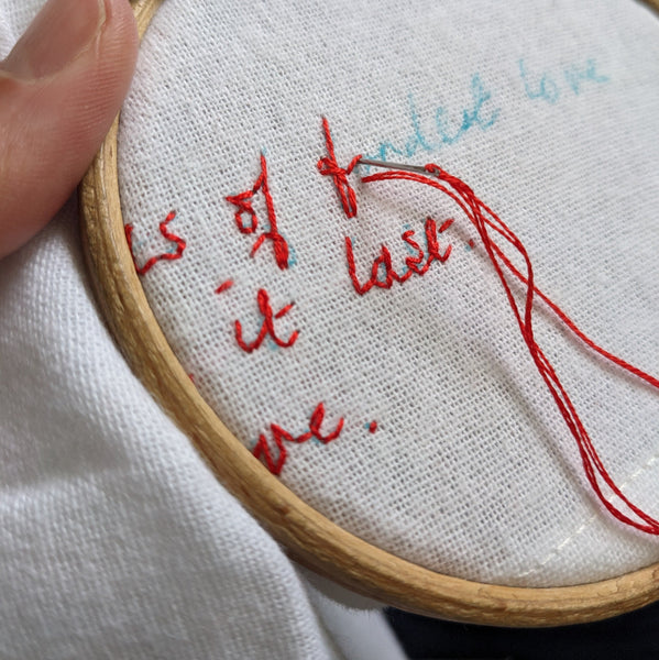 White cotton fabric that is in an embroidery hoop and being embroidered with red thread. The embroidery reads 'fondest love'.