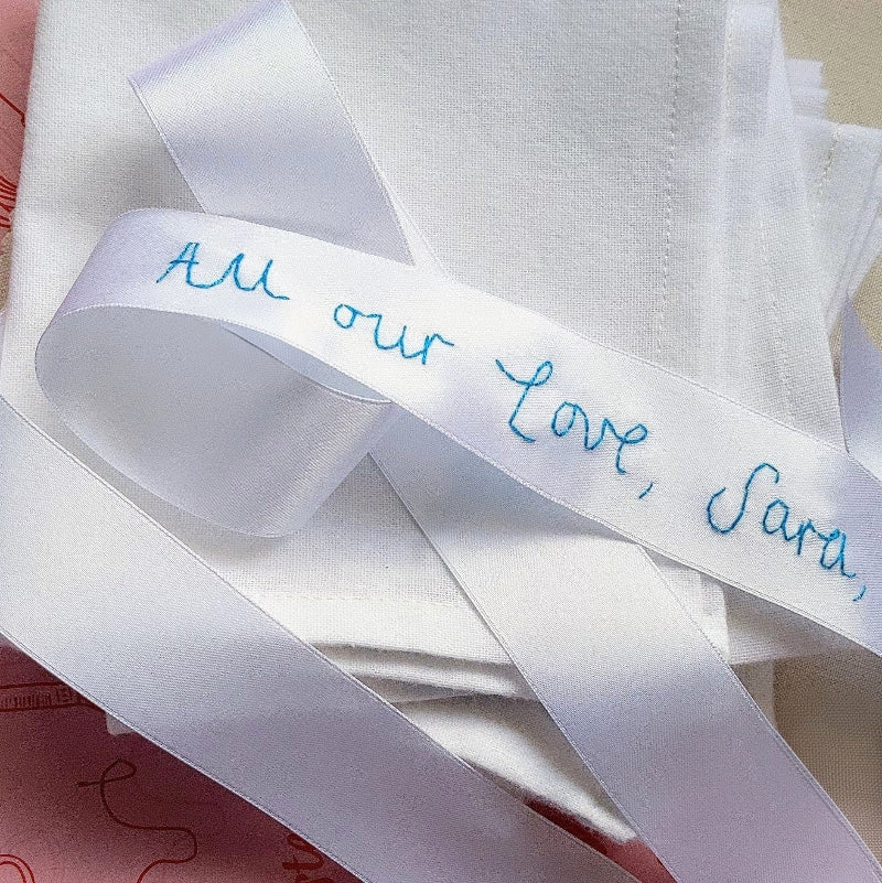 White wedding bouquet ribbon with blue handwriting embroidered on it that says 'All our love, Sara'