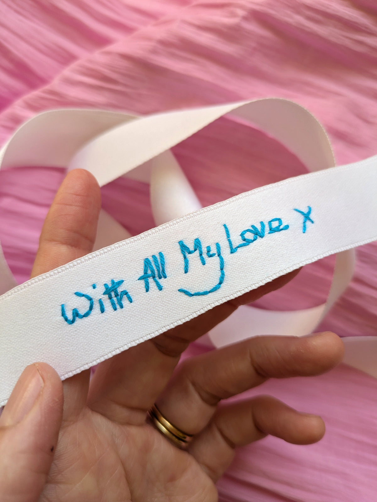 A hand holding a white ribbon with blue embroidered handwriting that reads 'With All My Love x' against a pink background.