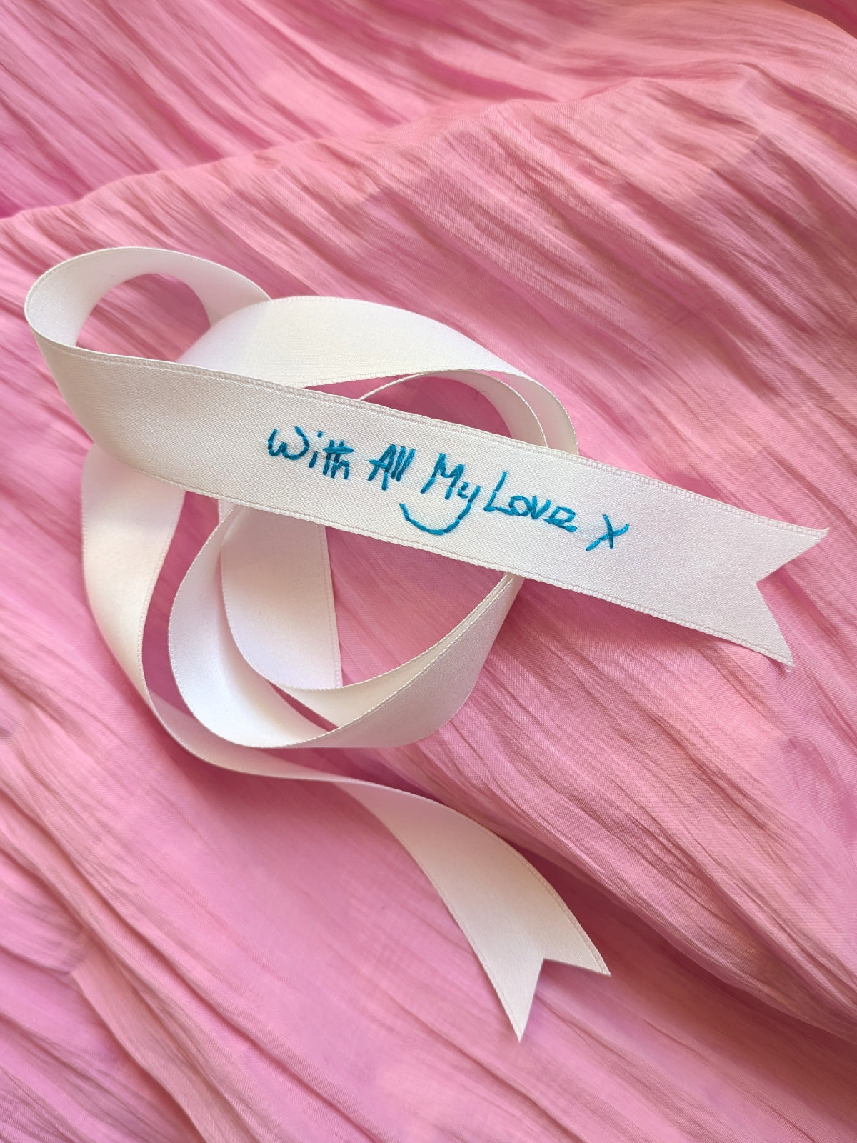 A white ribbon embroidered with blue handwriting reading 'With All My Love, X' resting on a textured pink fabric background.