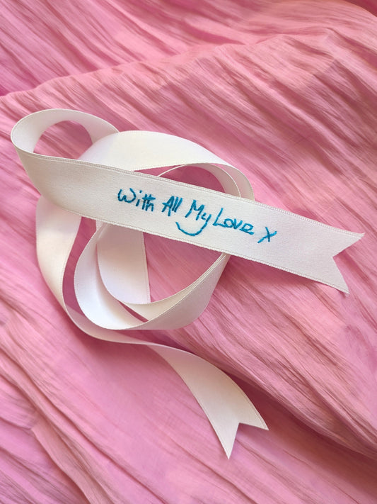 A white ribbon embroidered with blue handwriting reading 'With All My Love, X' resting on a textured pink fabric background.