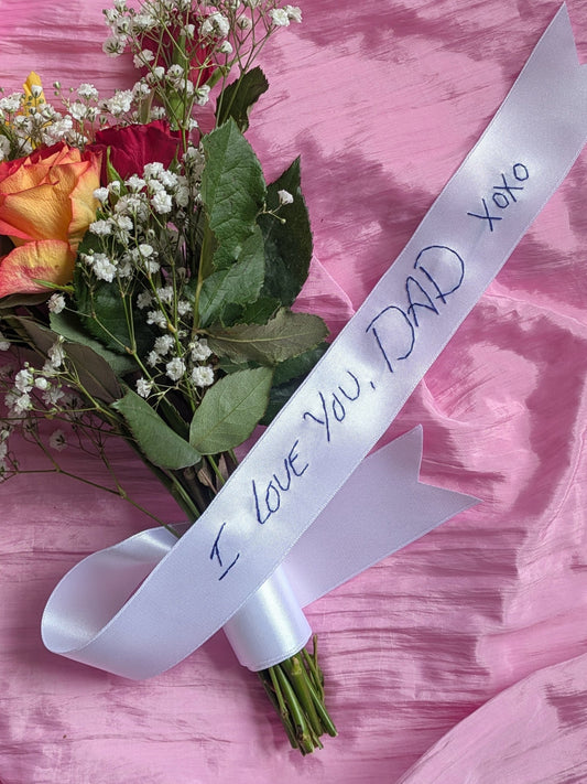 Bouquet of flowers with a white personalised handwriting wedding bouquet ribbon that reads 'I love you, Dad xoxo' on a pink background