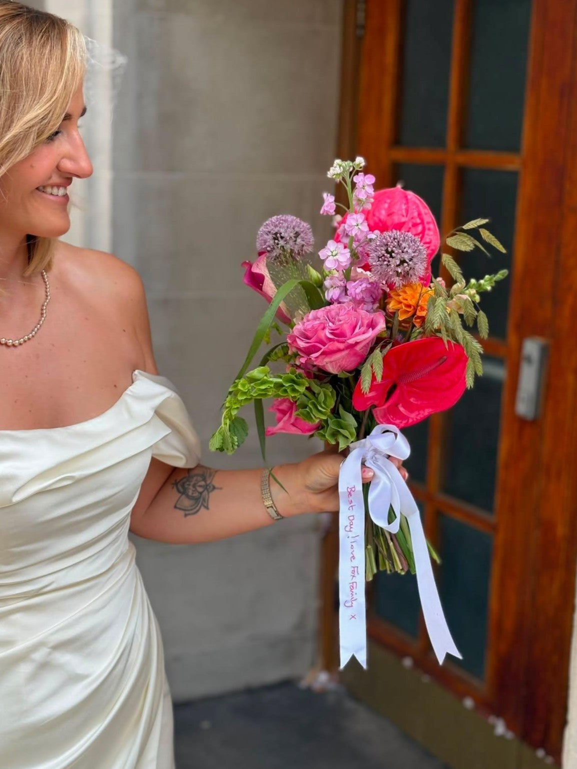 A woman in an off-the-shoulder white wedding dress holds a vibrant wildflower bouquet with pink roses, red anemones, purple thistles, and green foliage, tied with a white ribbon with embroidery.