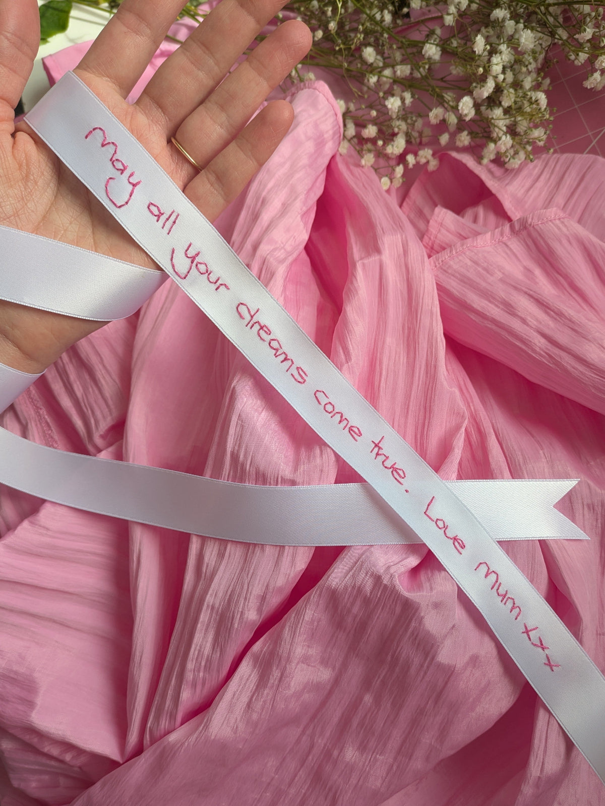 A white ribbon embroidered with a handwritten message, held by hands against a pink background.
