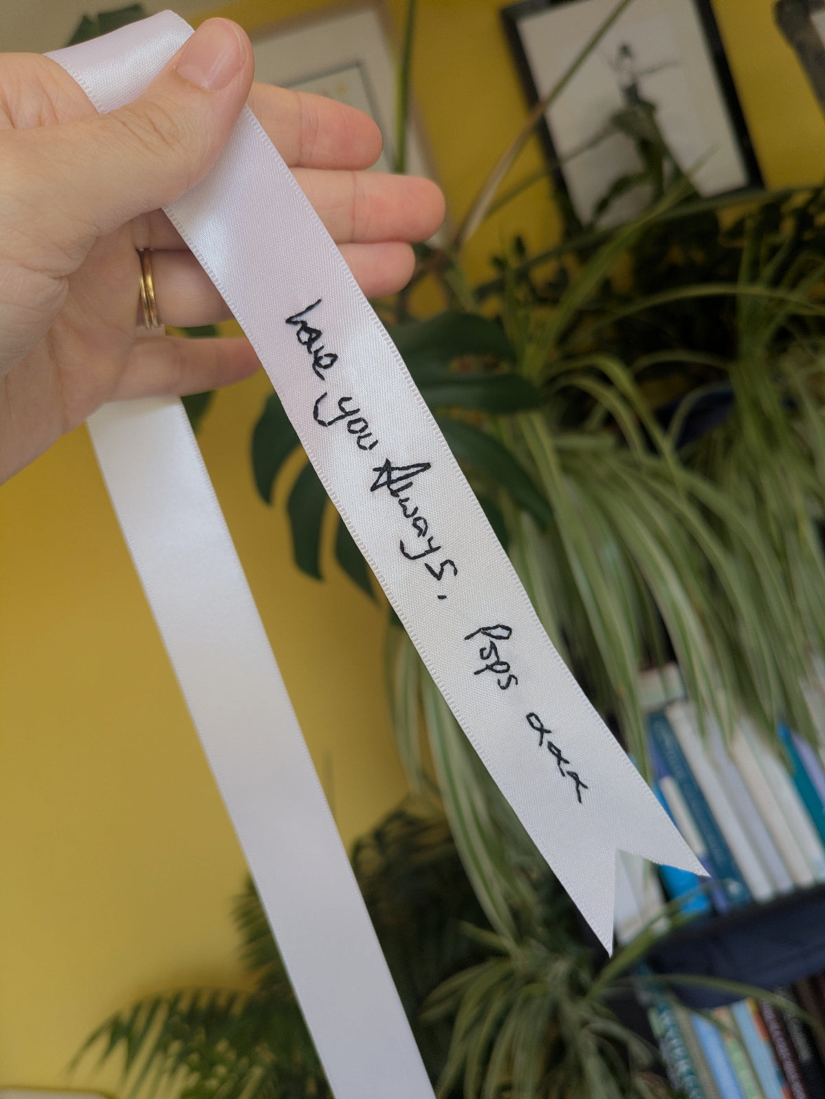 A hand holds a white ribbon with handwritten black embroidery reading 'love you Always, Pops are' against a yellow wall with houseplants in the background.