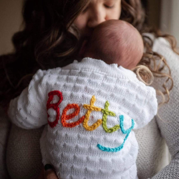 A woman is holding a baby wearing a white knitted cardigan with 'Betty' embroidered in colourful letters.