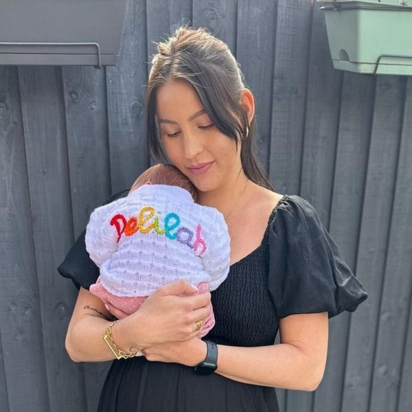 Woman holding a baby wearing a white knitted cardigan with the name 'Delilah' embroidered in colourful threads.