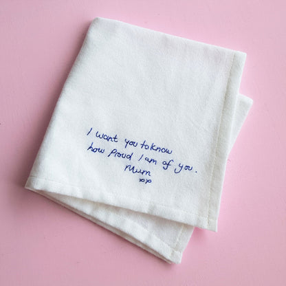 A white cotton handkerchief with navy embroidered handwriting that reads 'I want you to know how proud I am of you. Mum xx'