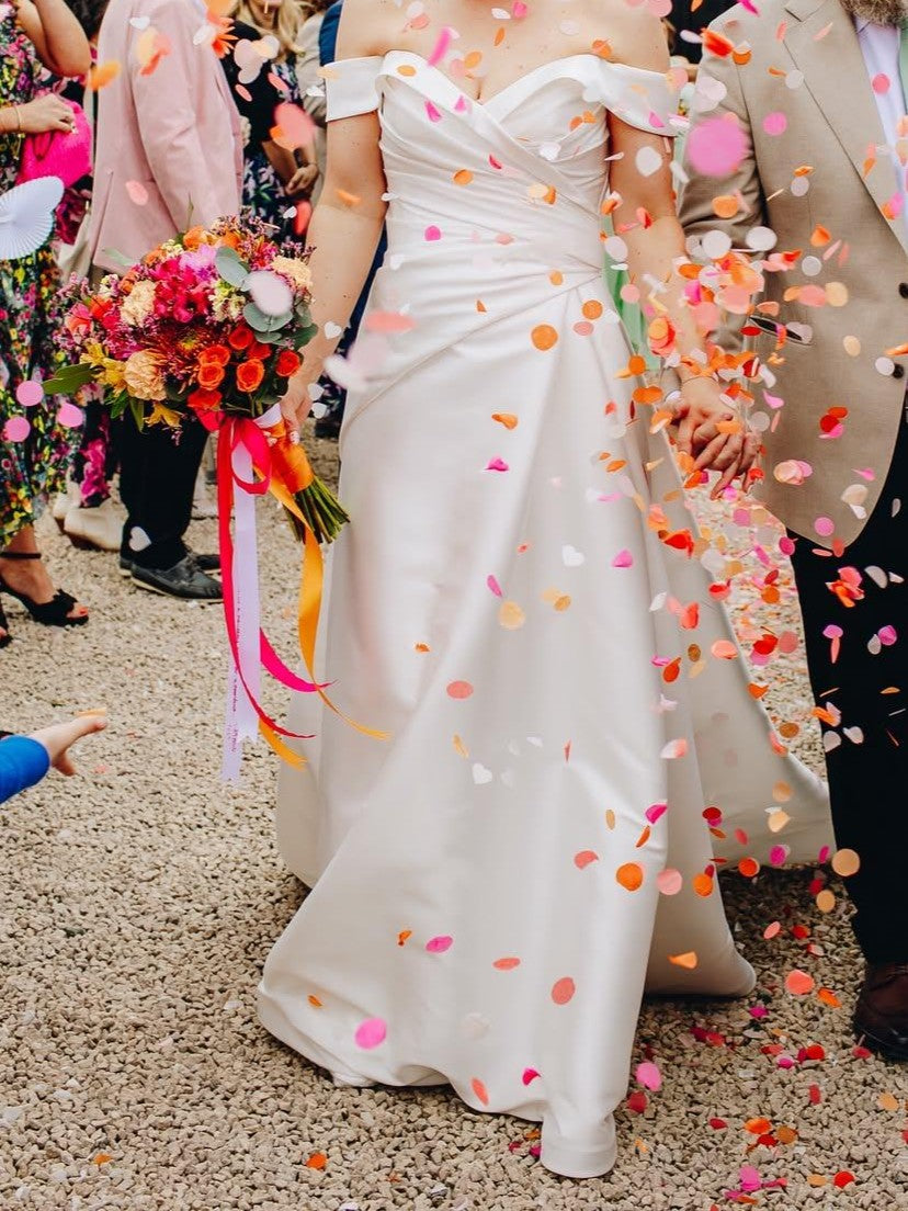 A woman in a white off-the-shoulder wedding gown walking outdoors while being showered with colourful confetti. She is holding a vibrant floral bouquet with pink and orange ribbons, surrounded by guests in a festive outdoor setting.