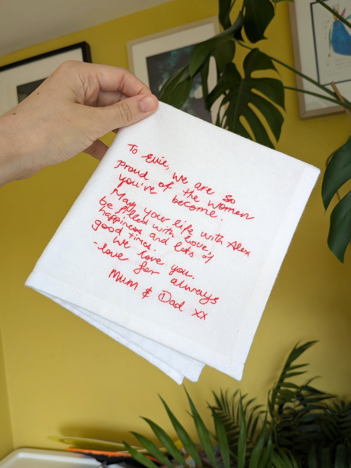 A hand holds a cotton handkerchief embroidered with a handwritten message, photographed indoors against a yellow wall with framed art and green plants visible in the background.