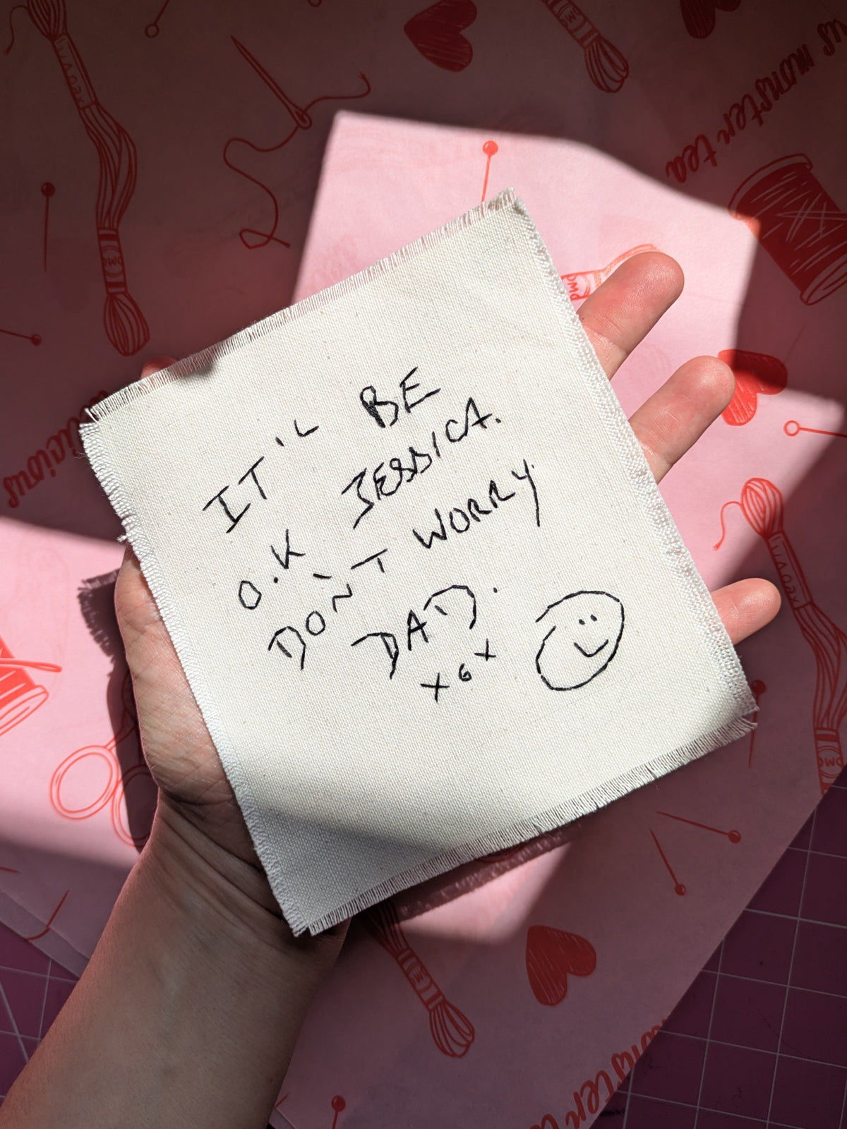 A hand holds a square off-white fabric patch with handwritten message that has been hand embroidered.