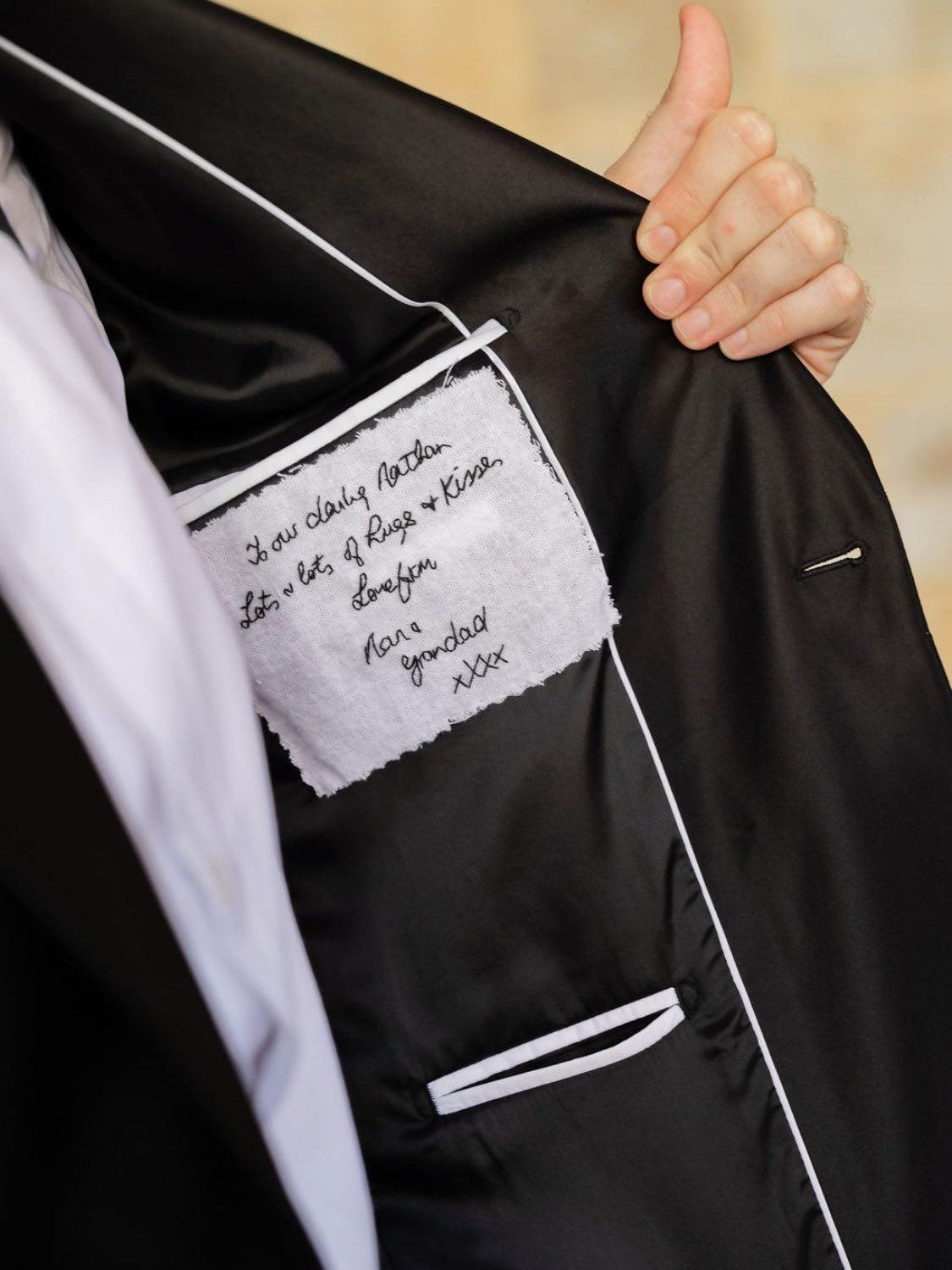 Close-up of a black suit jacket, opened to reveal a white patch embroidered with a handwritten message, with a hand holding the lapel open.