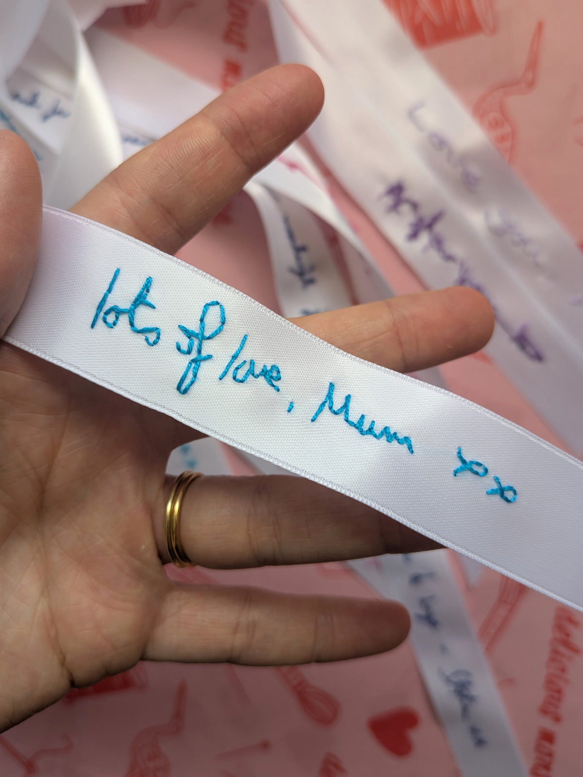 A hand holding a white satin ribbon embroidered with blue cursive handwriting that reads 'lots of love, Mum xx'. More signed white ribbons are visible in the background on a pink surface.
