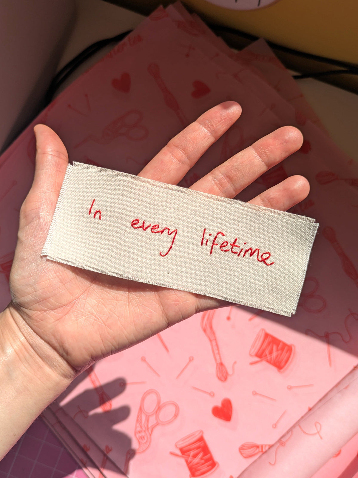 A hand holding a rectangular fabric patch with red embroidered text reading 'In every lifetime'. Pink patterned fabric is visible in the background.