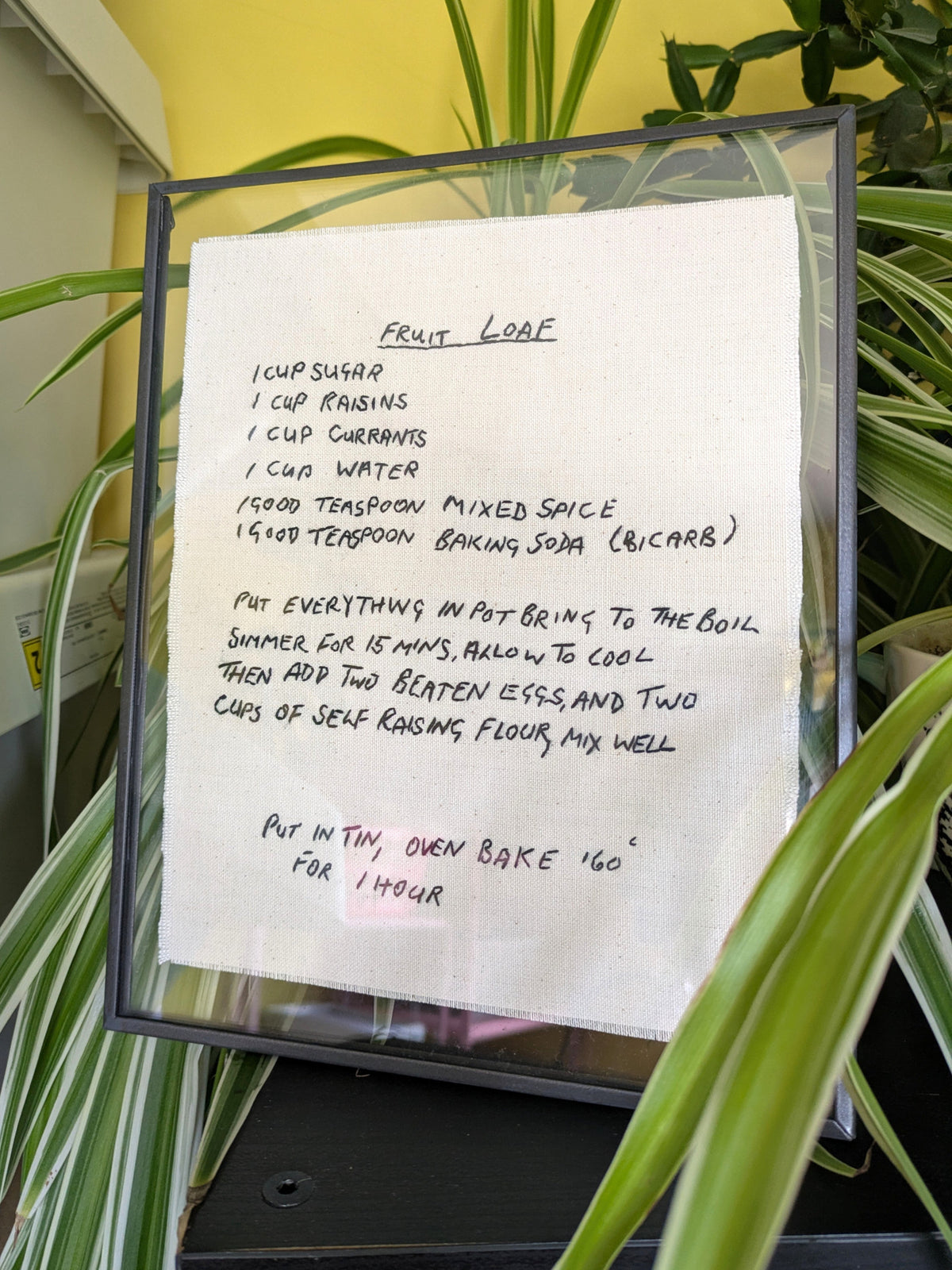 A framed handwritten recipe for fruit loaf, hand embroidered on cotton fabric and displayed in a glass-front frame among green houseplant leaves.