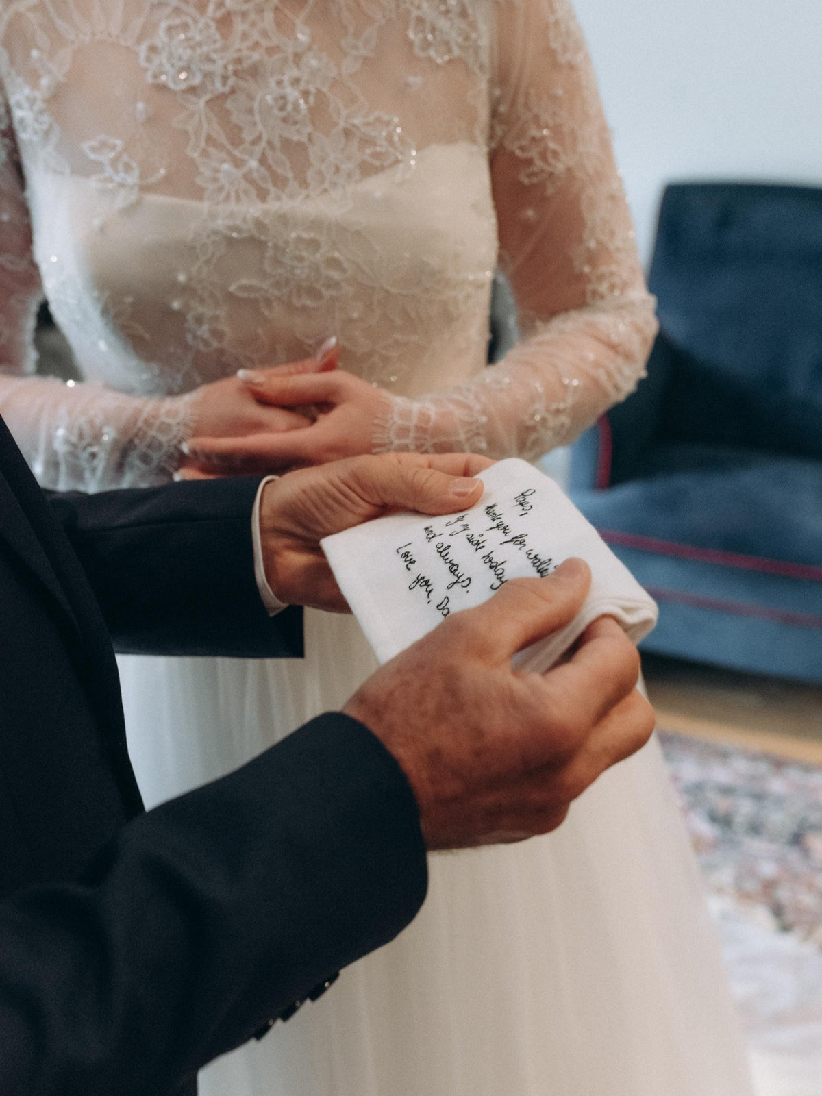 A close-up lifestyle shot of a bride in a cream lace wedding gown next to the father of the bride who is holding a handkerchief embroidered with a handwritten message.