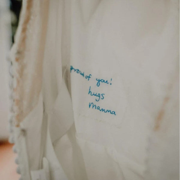A white cotton patch that has been sewn into the lining of a wedding dress. It is embroidered in blue thread with handwriting that reads 'proud of you! hugs Mamma'