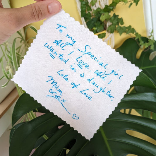 A white cotton wedding dress label with blue embroidered handwriting that reads 'To my special girl, all I love, all I wanted in a daughter. Lots of love Mum xxx'