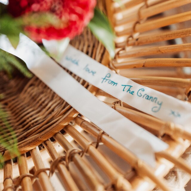 A white satin ribbon is tied around a wedding bouquet of flowers. There is a handwritten message embroidered on the ribbon that read 'with love from The Granny xx' in blue thread.