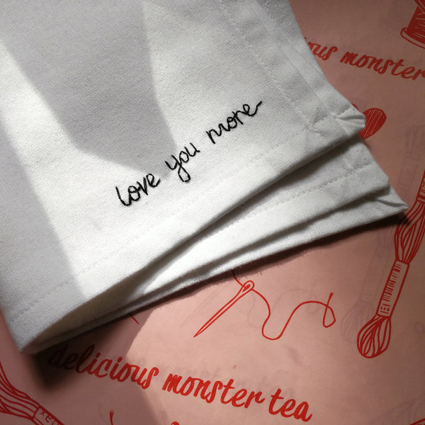 White hanky with 'love you more' embroidery on a pink background with red text and graphics.