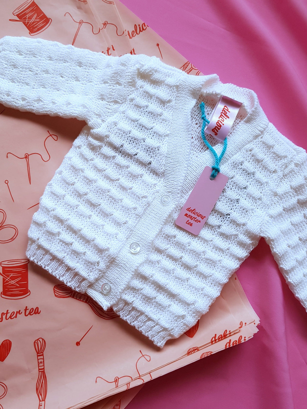 White knitted baby cardigan with branded swing tag laid flat on pink tissue paper