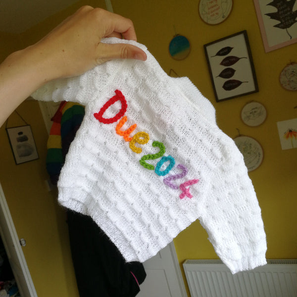 White knitted baby cardigan with 'Due 2024' embroidered on it in colourful letters held by a hand against a yellow wall.
