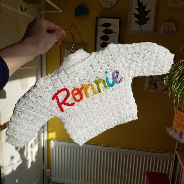 White knitted baby cardigan with 'Ronnie' embroidered in colourful letters on a hanger.
