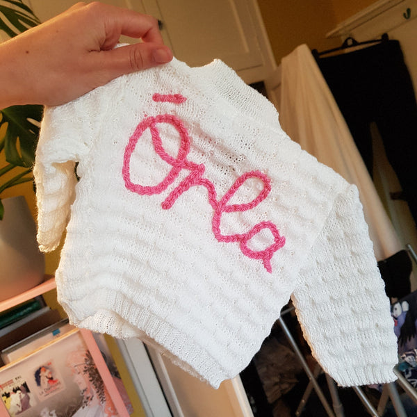 White baby cardigan with pink embroidered name 'Orla' held by a hand in a casual indoor setting.