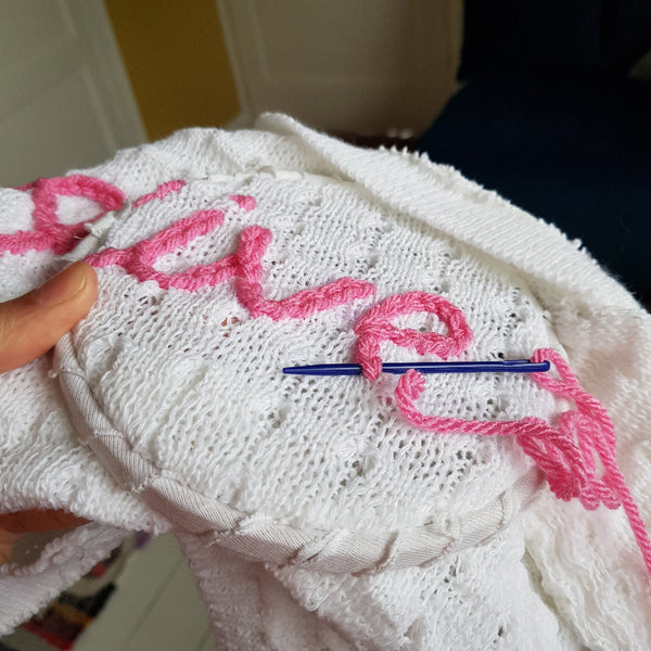 White baby cardigan with pink embroidered text and a blue needle, held by a hand.