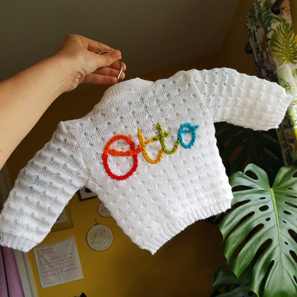 White knitted baby cardigan with 'Otto' embroidered in colourful letters, held by a hand.