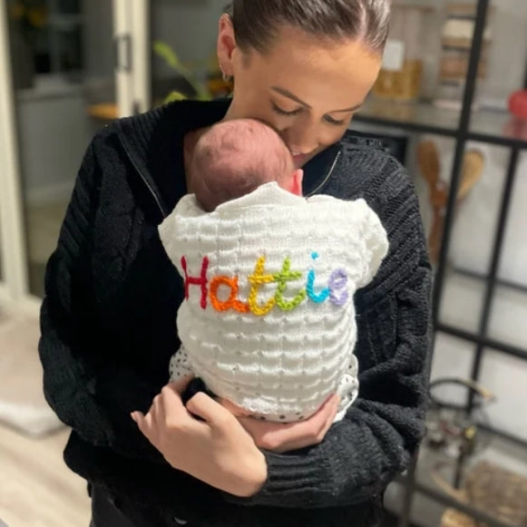 Woman holding a baby wearing a white knitted cardigan with 'Hattie' embroidered in colourful letters.
