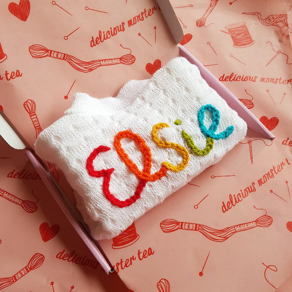 Embroidered baby cardigan with colourful letters on a pink patterned background.