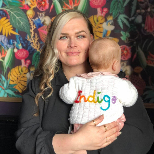 Woman holding a baby wearing a white sweater with colorful text in front of a colorful wall.