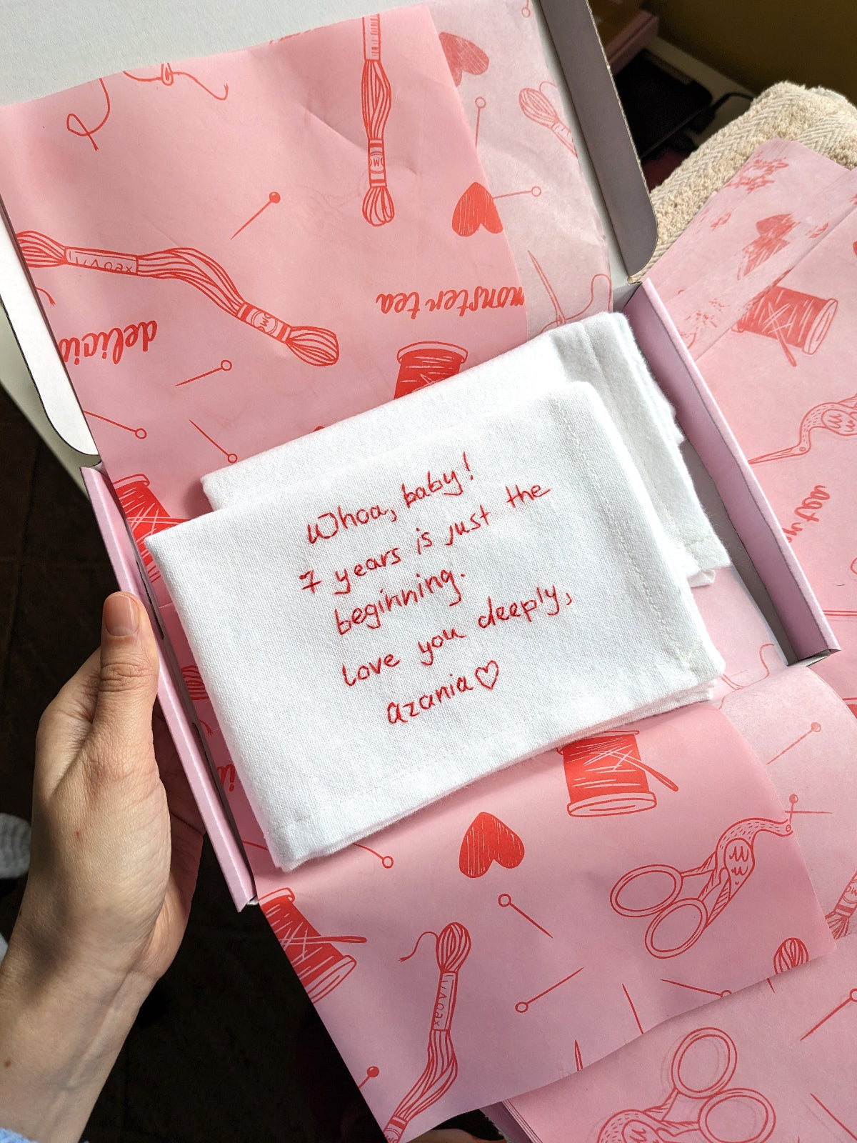 A hand holds an open pink gift box lined with pink sewing-themed tissue paper and a folded white handkerchief embroidered with red handwritten text that reads, 'Whoa, baby! 2 years is just the beginning. Love you deeply, Azonia <3'.